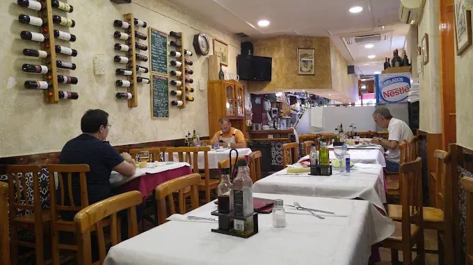 Interior de restaurante con mesas preparadas; gente comiendo. Botelleros en la pared.