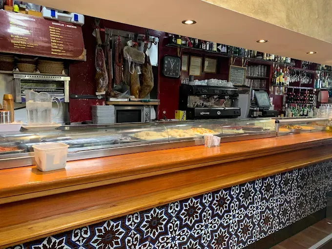 Interior de bar con mostrador de madera y azulejos, exposición de alimentos y embutidos colgantes.