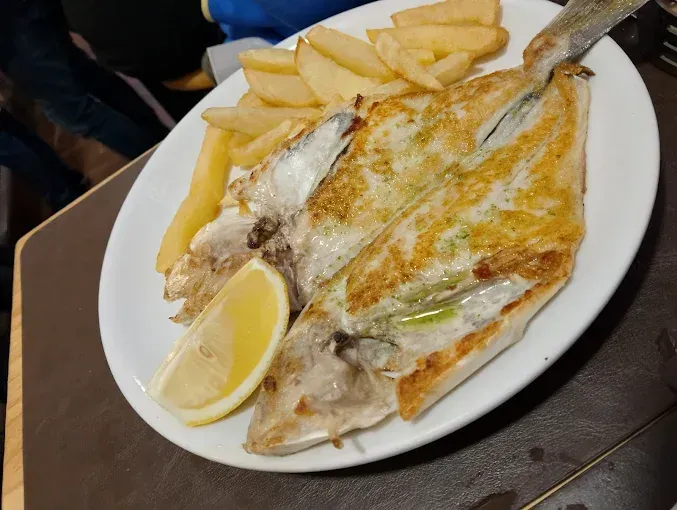 Pescado a la parrilla con patatas fritas y rodaja de limón en un plato blanco.