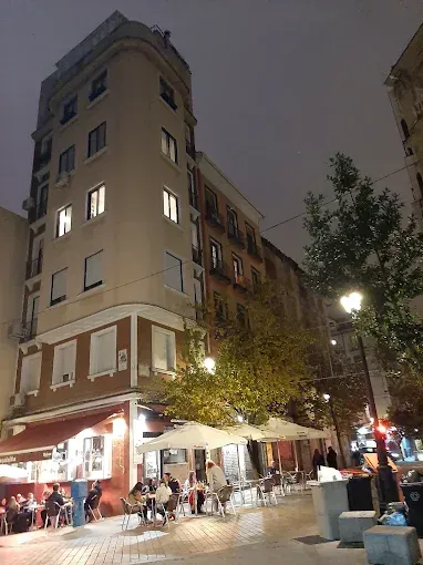 Escena callejera nocturna con edificios altos, asientos en cafeterías y gente cenando al aire libre.