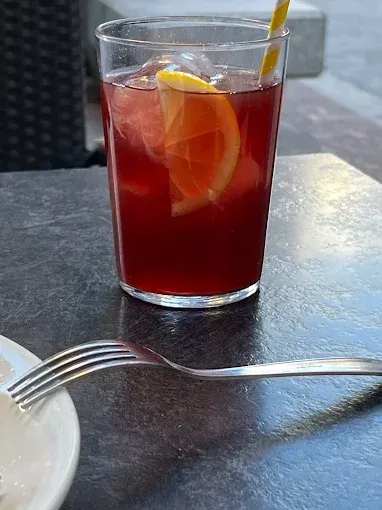 Vaso de bebida roja con hielo y rodaja de limón, junto a un tenedor y un plato sobre una mesa.
