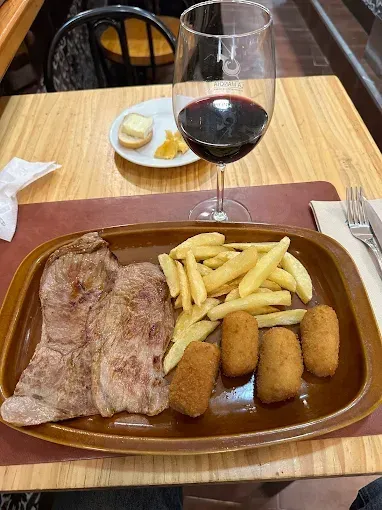 Cena de bistec con patatas fritas y croquetas, copa de vino y pequeño plato de queso sobre mesa de madera.