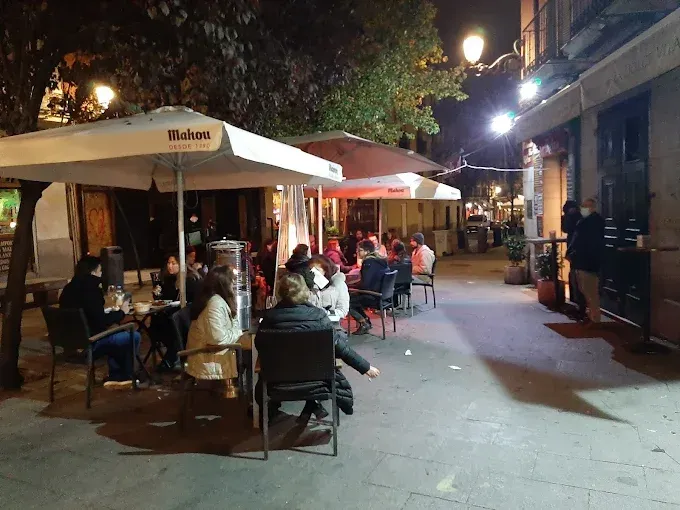 Cafetería al aire libre con gente sentada en mesas bajo sombrillas por la noche. Las farolas iluminan la zona.