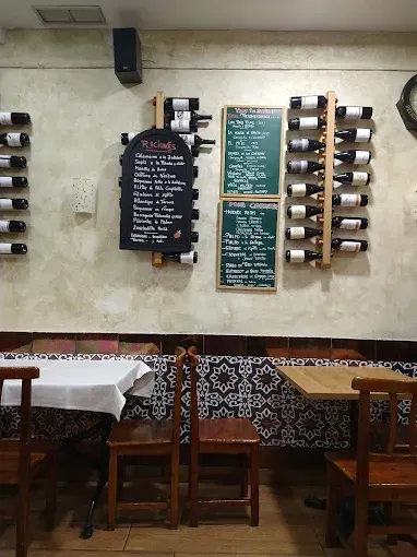 Interior de restaurante con botellas de vino en la pared, tableros de menú y mesas y sillas de madera.