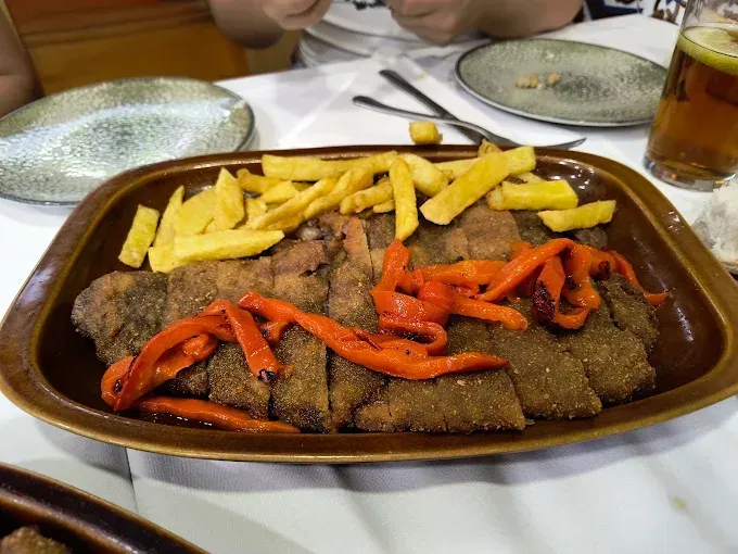 Bandeja de carne frita con patatas fritas y pimientos rojos, sobre mantel blanco, ambientación de restaurante.