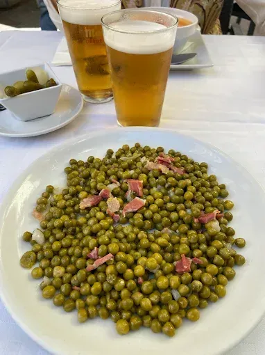 Plato de guisantes con jamón, vasos de cerveza y guarniciones sobre mantel blanco.