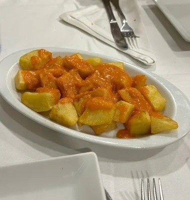 Patatas bravas: patatas fritas con salsa picante roja en un plato ovalado blanco. Tenedores y cuchillos cerca.