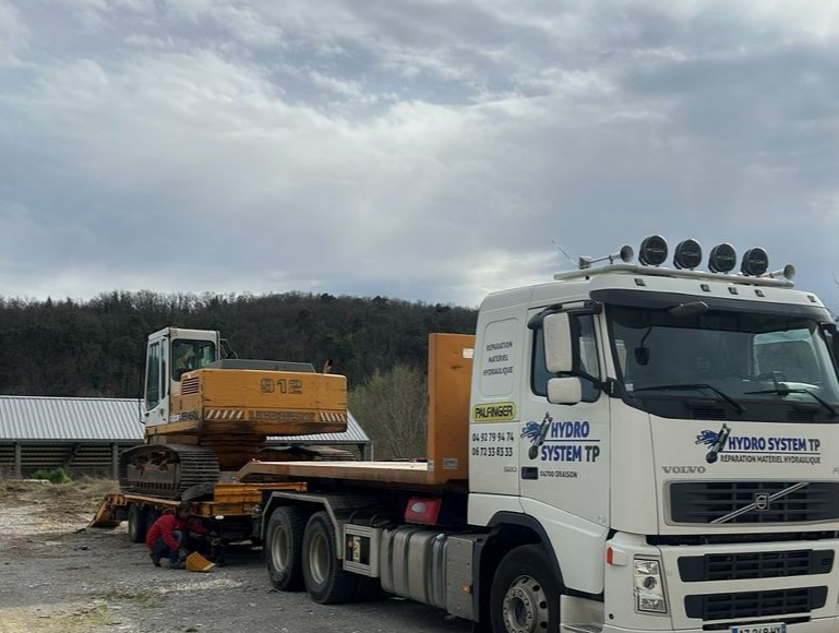 Le camion d'HYDRO SYSTEM TP transporte une excavatrice jaune au bord d'une route de campagne.