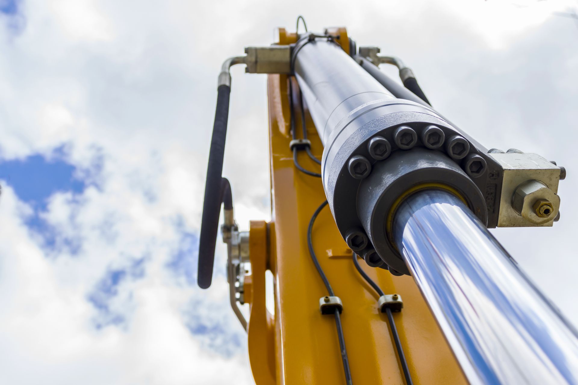 Un bras hydraulique jaune sur fond de ciel bleu, avec un piston chromé brillant déployé.