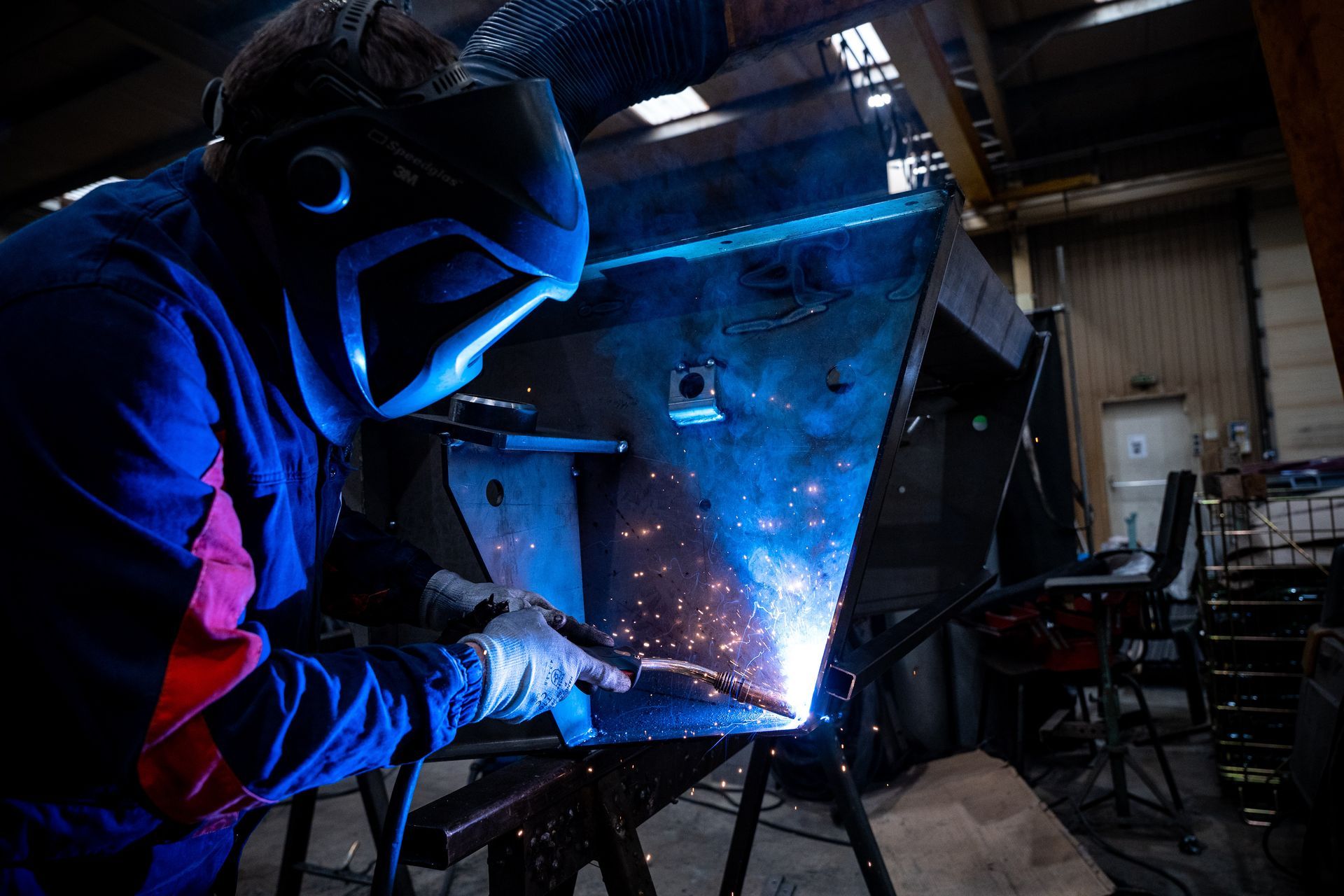Un soudeur en tenue de protection bleue soude des pièces métalliques brillantes dans un atelier.