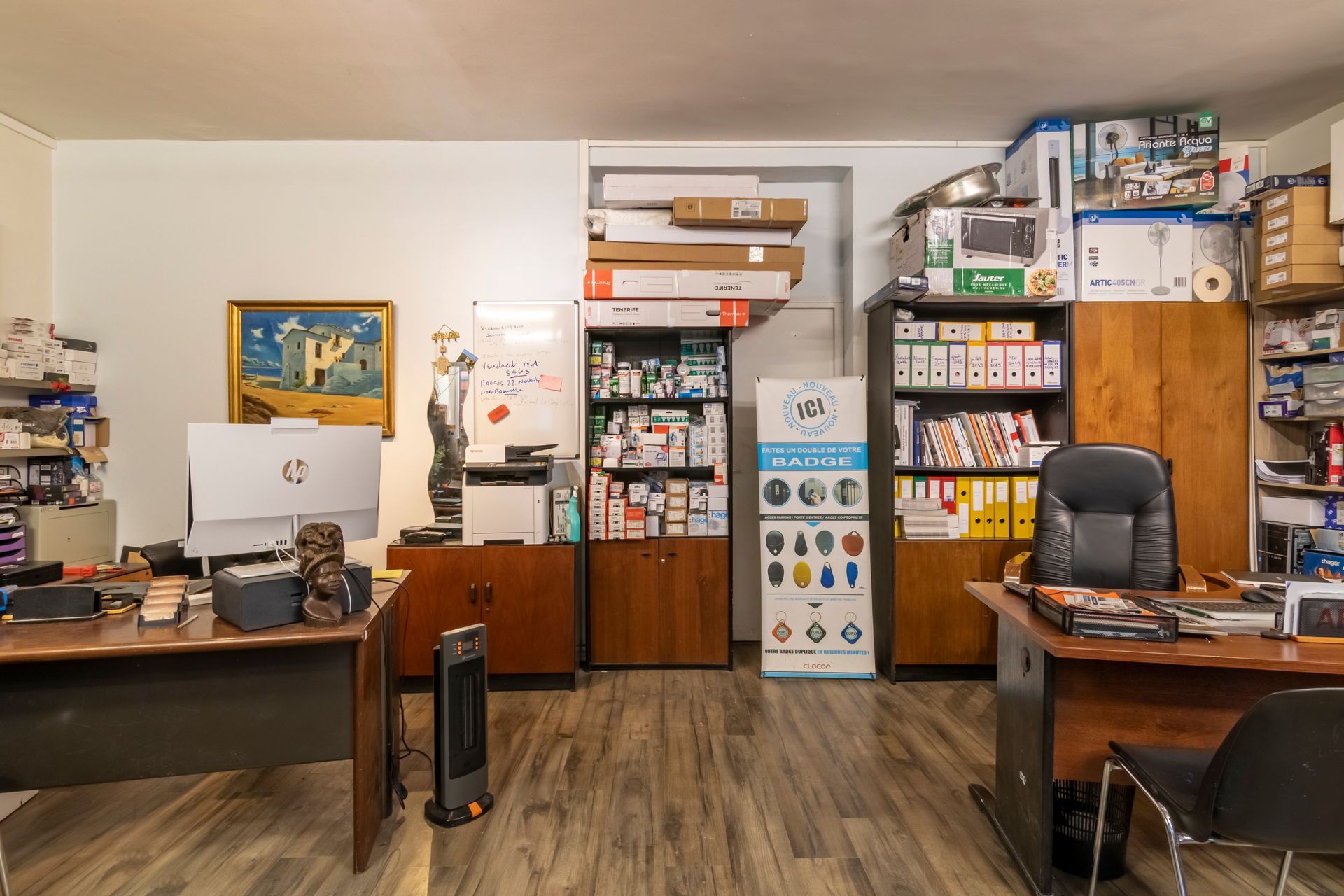 Pièces avec 2 bureaux et matériel informatique dessus, des armoires remplies de boîtes sont présentes au fond de la pièce