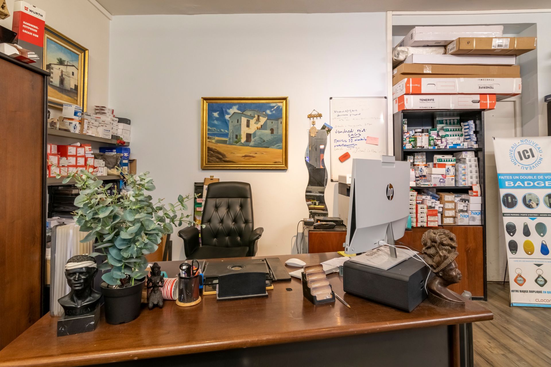 Un bureau encombré avec un ordinateur, des tableaux encadrés, des étagères et des plantes dans un petit espace de travail