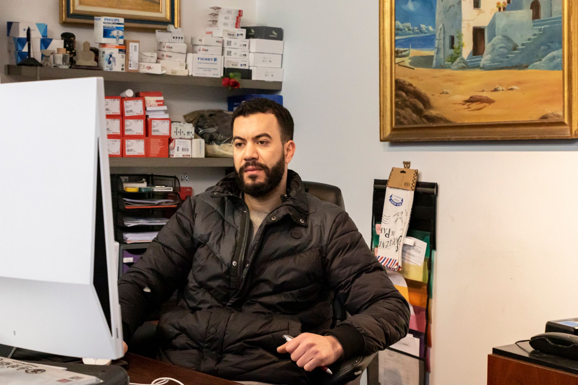 Un homme assis derrière un bureau, vêtu d'une veste noire, avec des étagères et des œuvres d'art encadrées derrière lui