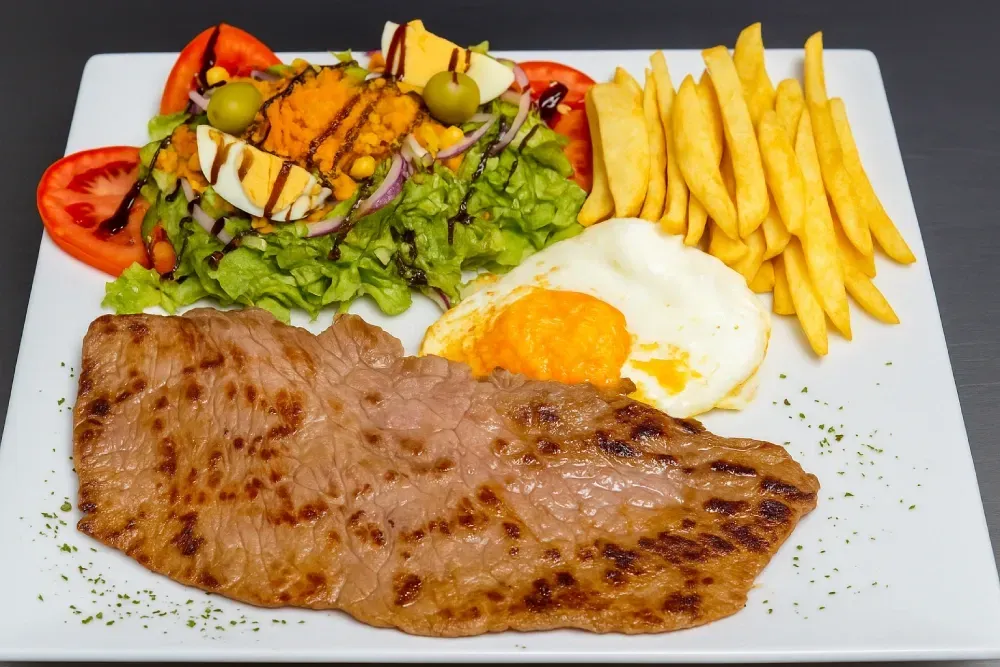 Bistec a la parrilla con huevo frito, patatas fritas y ensalada en un plato blanco.