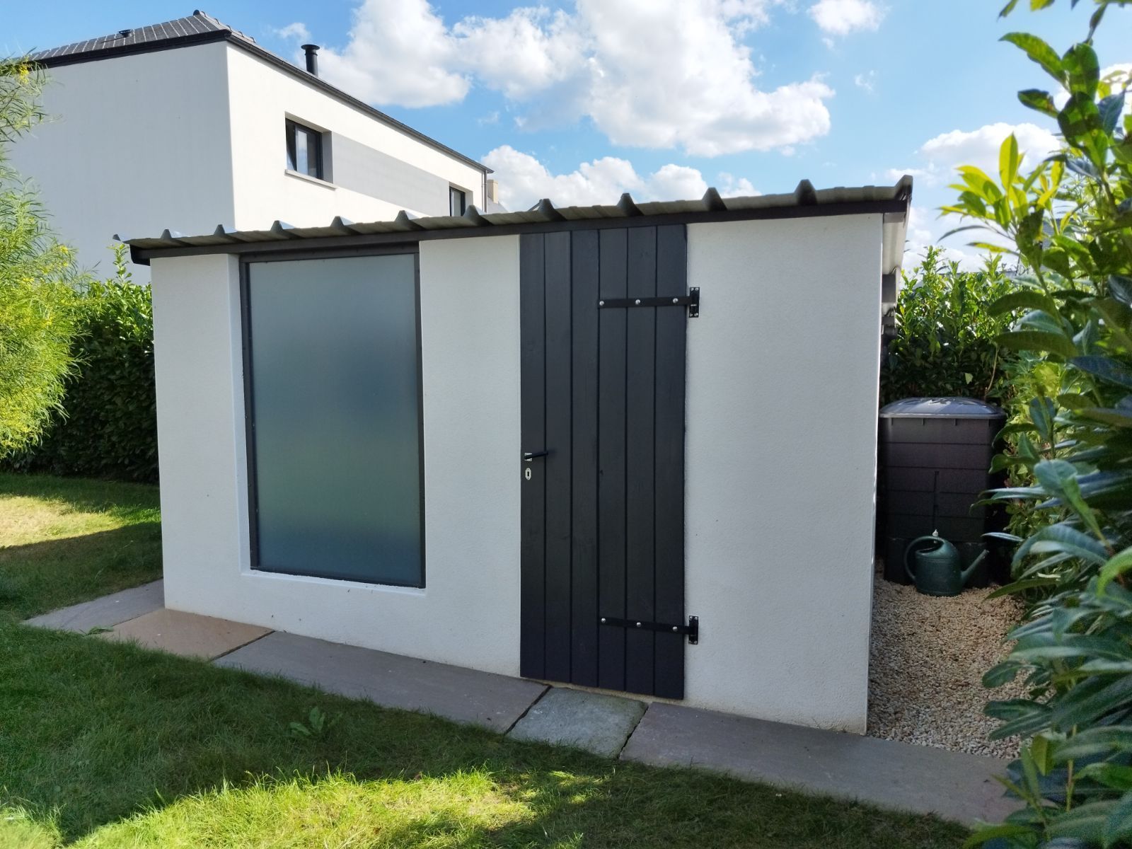 Petit abri de jardin blanc avec une porte grise et une fenêtre dépolie, jouxtant une maison et une pelouse.