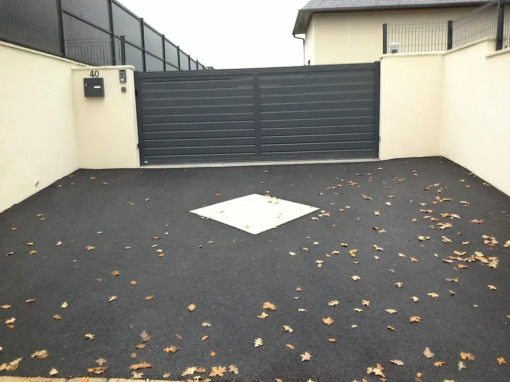 Cour pavée vide avec un portail sombre, des murs beiges, des feuilles éparses et une plaque d'égout carrée blanche.