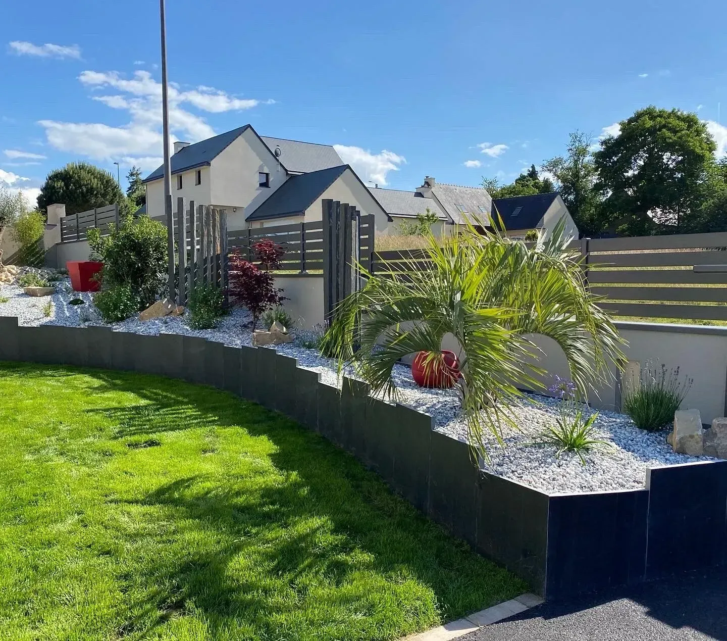 Un parterre de jardin paysager incurvé avec des rochers blancs, des palmiers et une clôture grise, jouxtant une pelouse verte et des maisons.