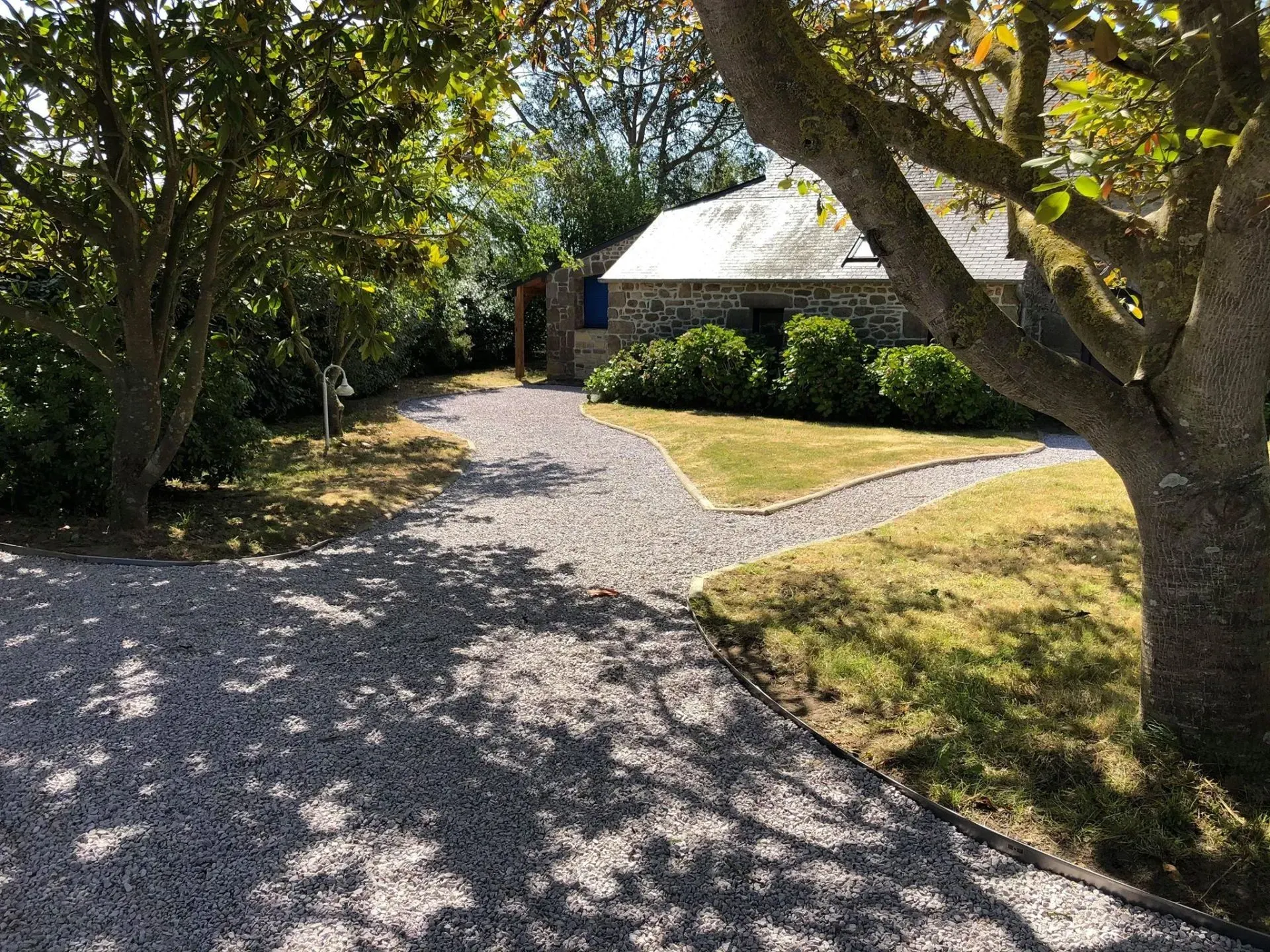Allée de gravier menant à une maison entourée d'arbres et baignée de soleil.