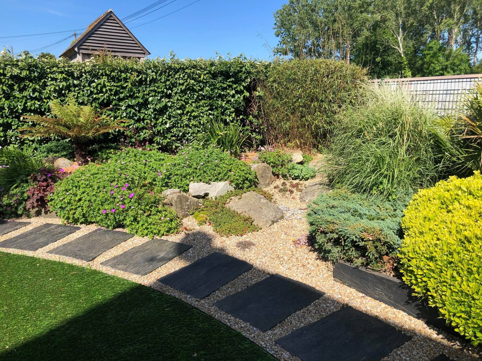 Jardin ensoleillé avec haies taillées, allée de pierres, plantes fleuries et pelouse verte.