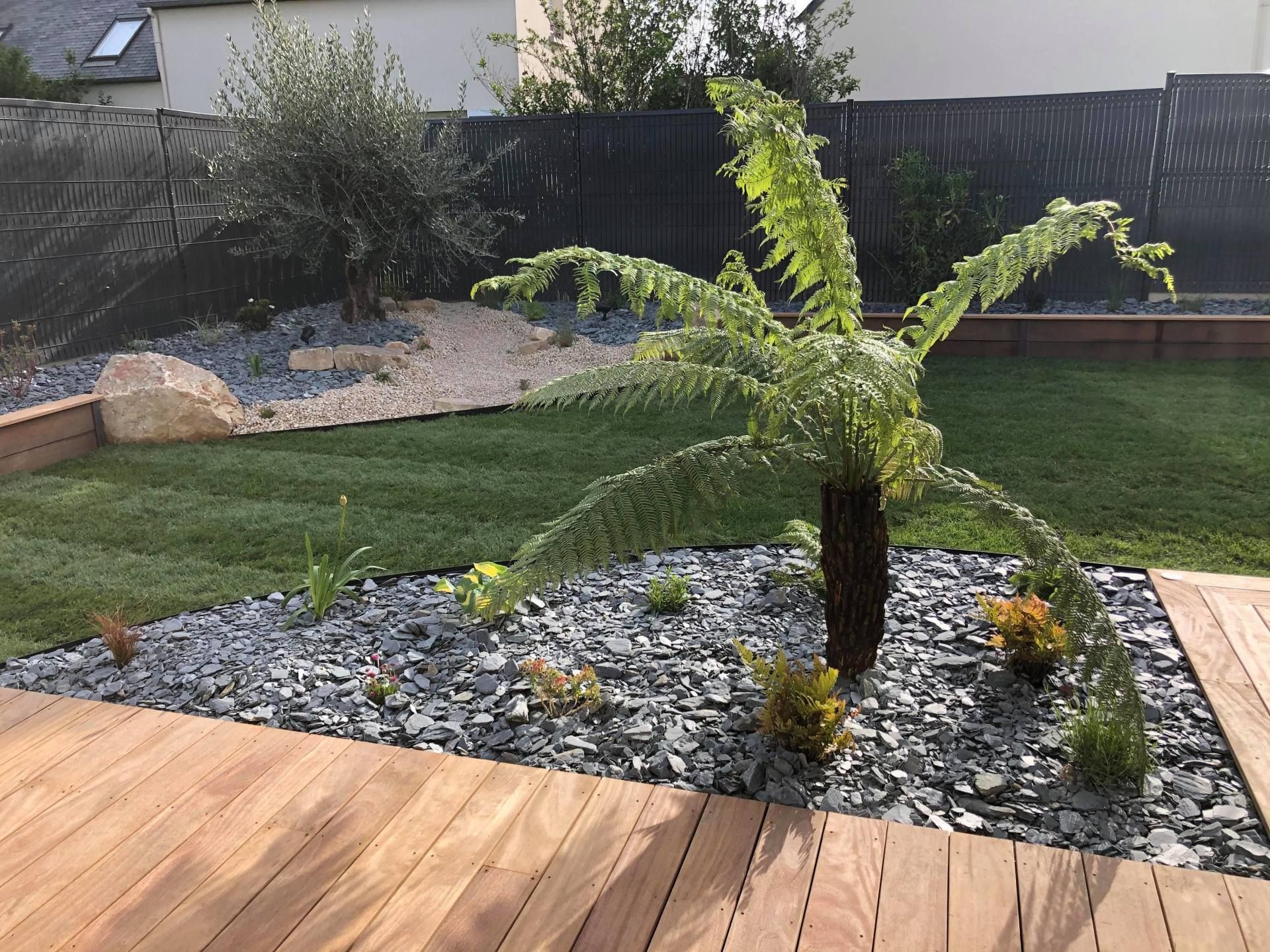 Jardin avec une plante ressemblant à un palmier dans un massif de pierres, bordé de pelouse et d'une terrasse en bois ; maisons en arrière-plan.