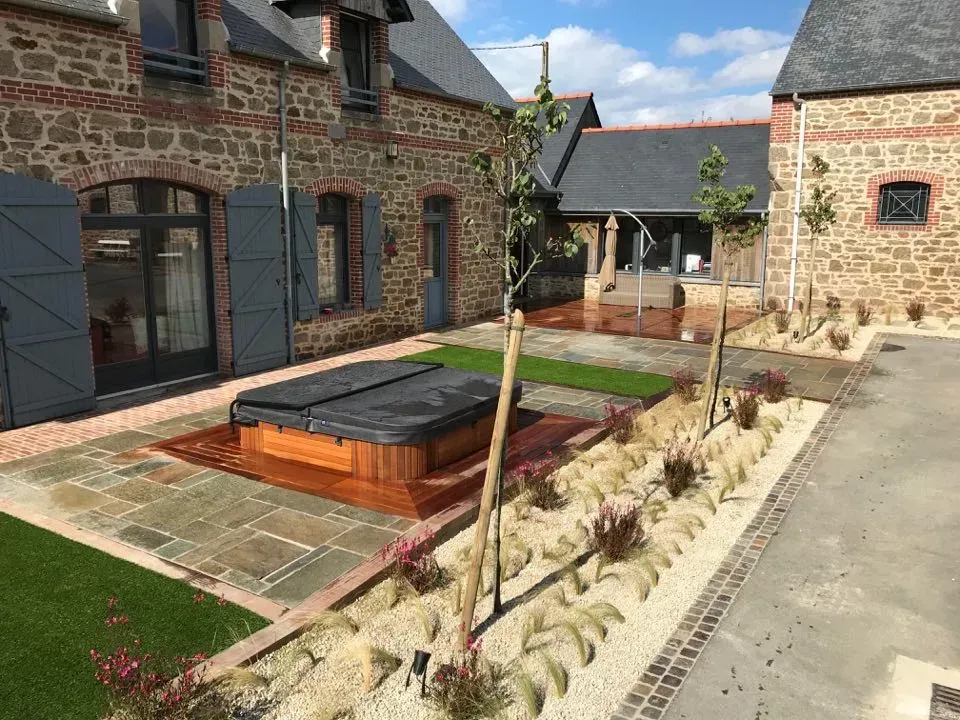 Cour intérieure avec bâtiments en pierre, jacuzzi surélevé en bois, pavés et parterres de fleurs aménagés.
