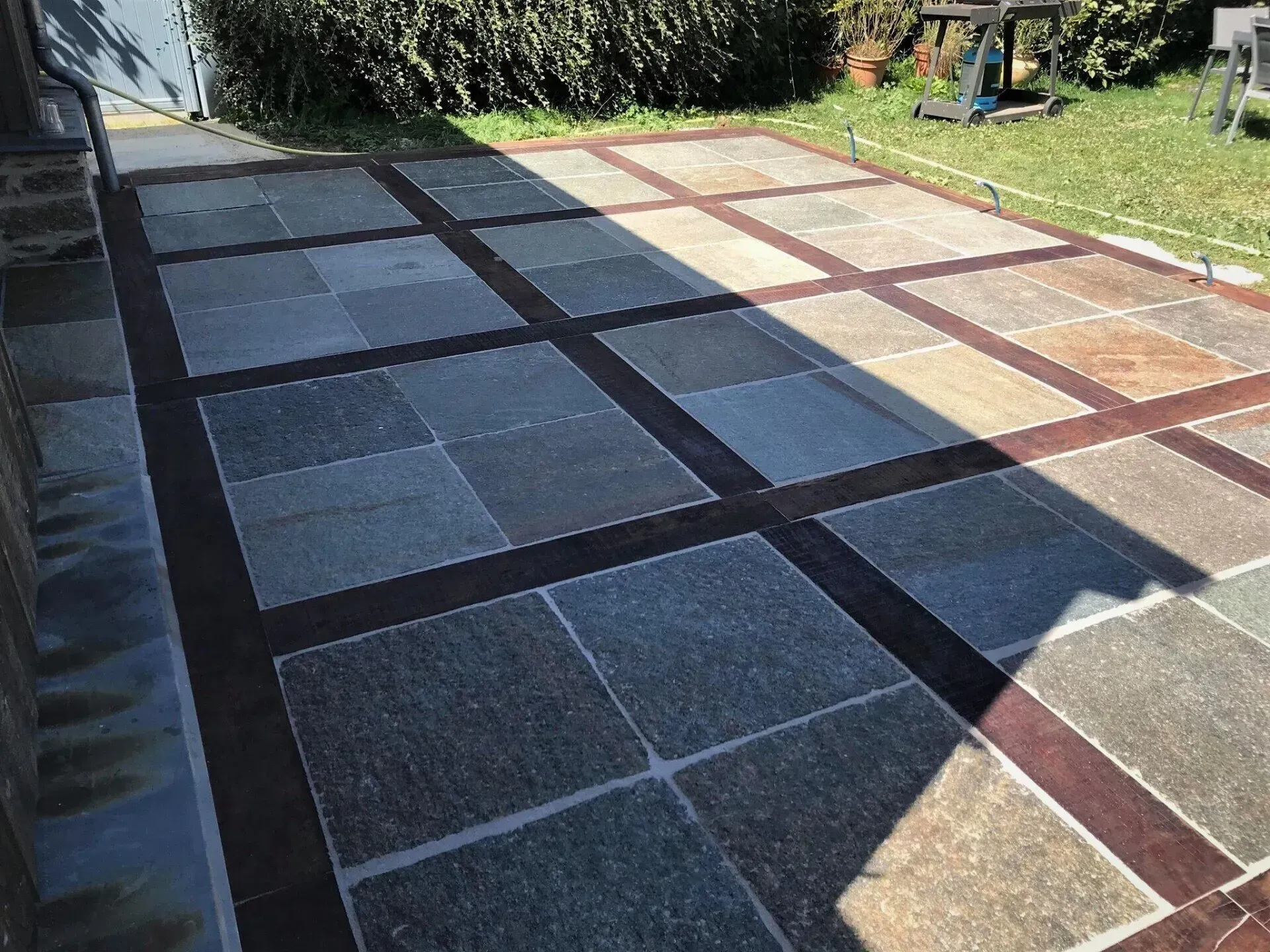 Terrasse extérieure ensoleillée avec de grands carreaux carrés à motifs géométriques gris, beiges et rouges.