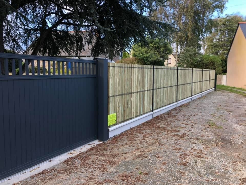Un grand portail noir jouxte une longue clôture en bois le long d'une allée de gravier, sous des arbres.