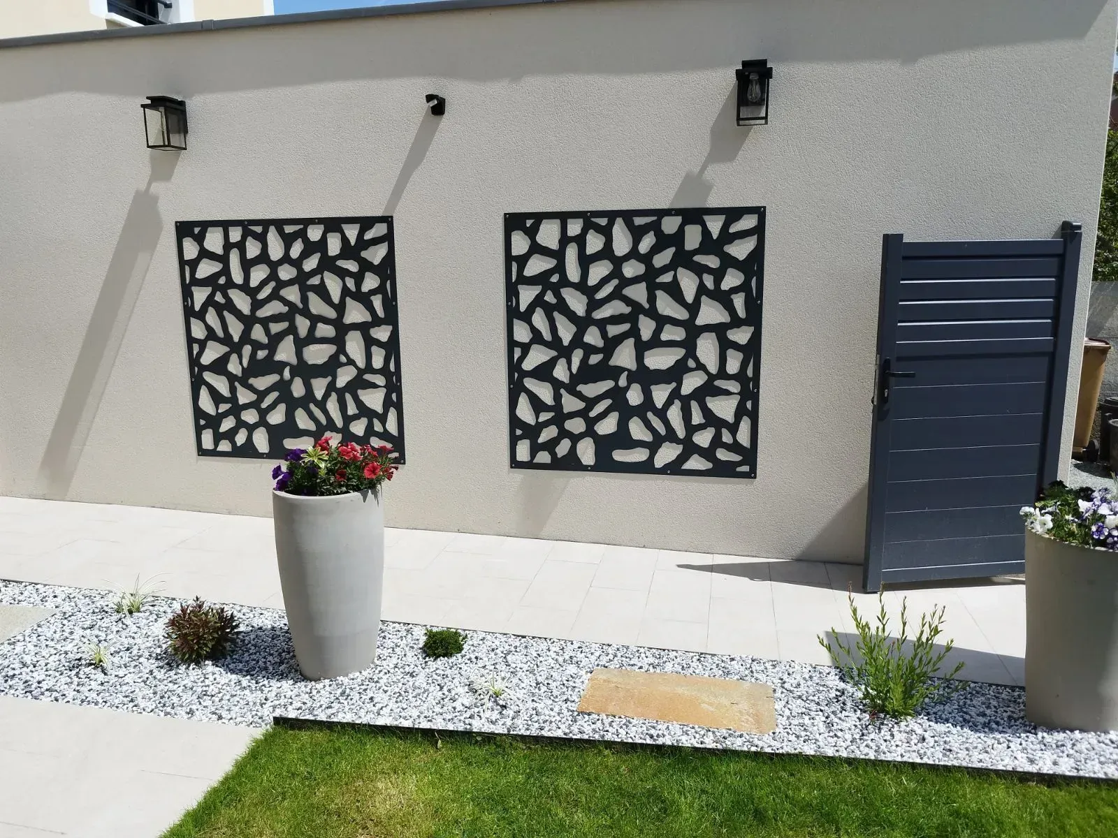 Extérieur de maison moderne avec panneaux muraux décoratifs, fleurs en pot et aménagement paysager en gravier.