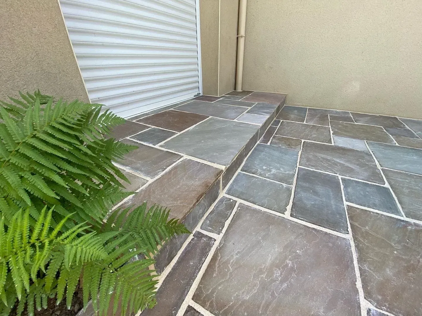 Coin de terrasse en pierre avec pavés gris, mur beige, volet roulant et fougère verte au premier plan.