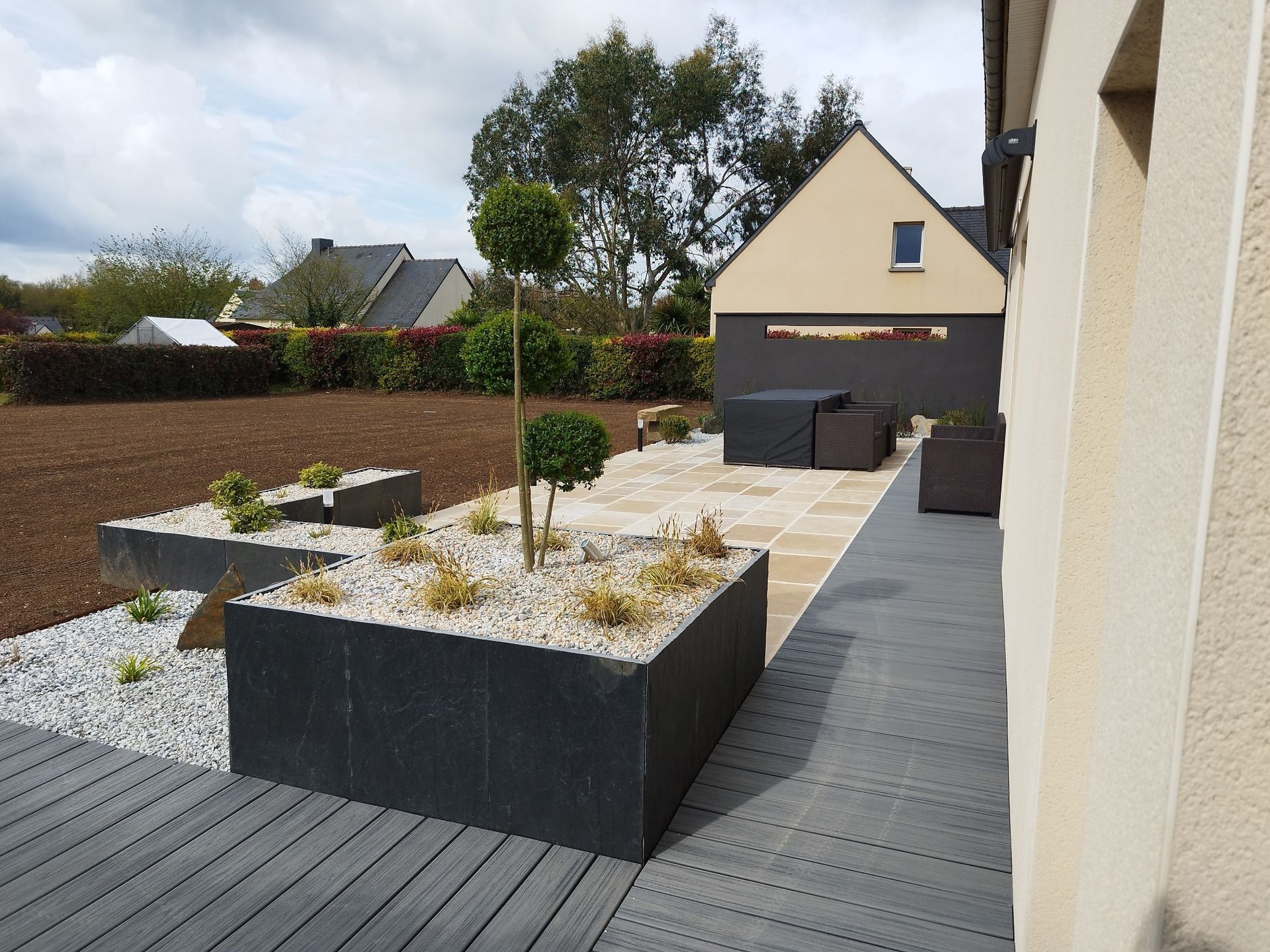Cour intérieure moderne avec jardinières noires surélevées, gravier, terrasse pavée et maison en arrière-plan.