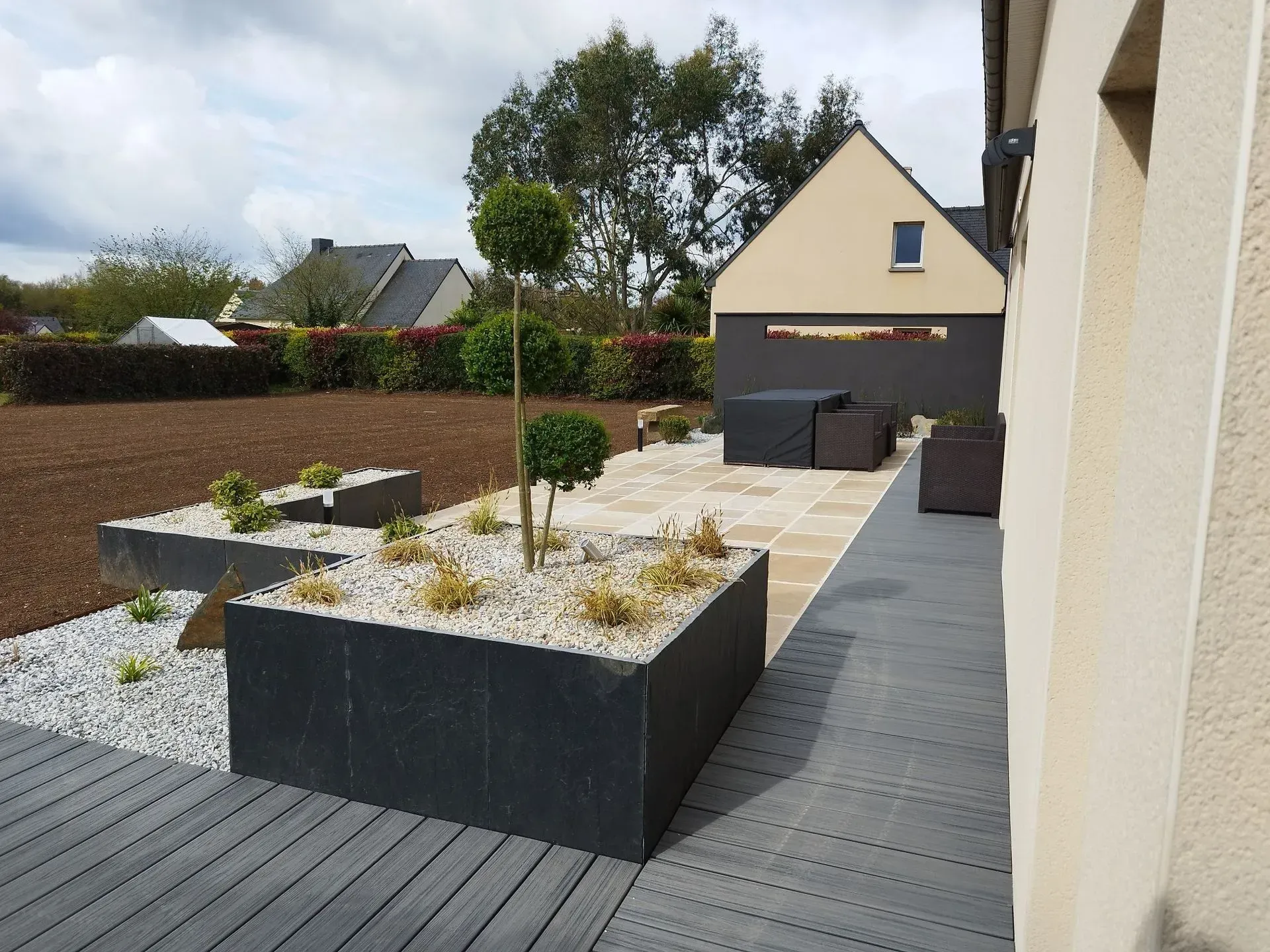 Cour intérieure moderne avec jardinières noires surélevées, parterres de gravier et allée pavée longeant une maison beige.
