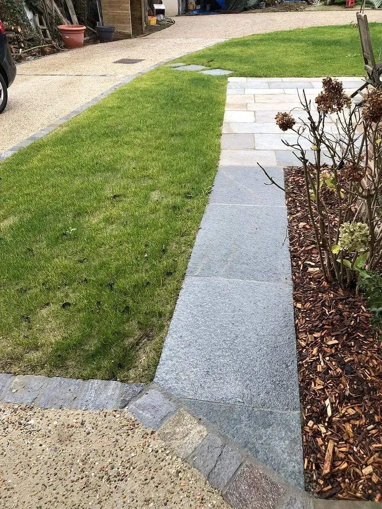 Allée en pierre incurvée longeant une pelouse verte et un parterre de fleurs paillé dans une cour.