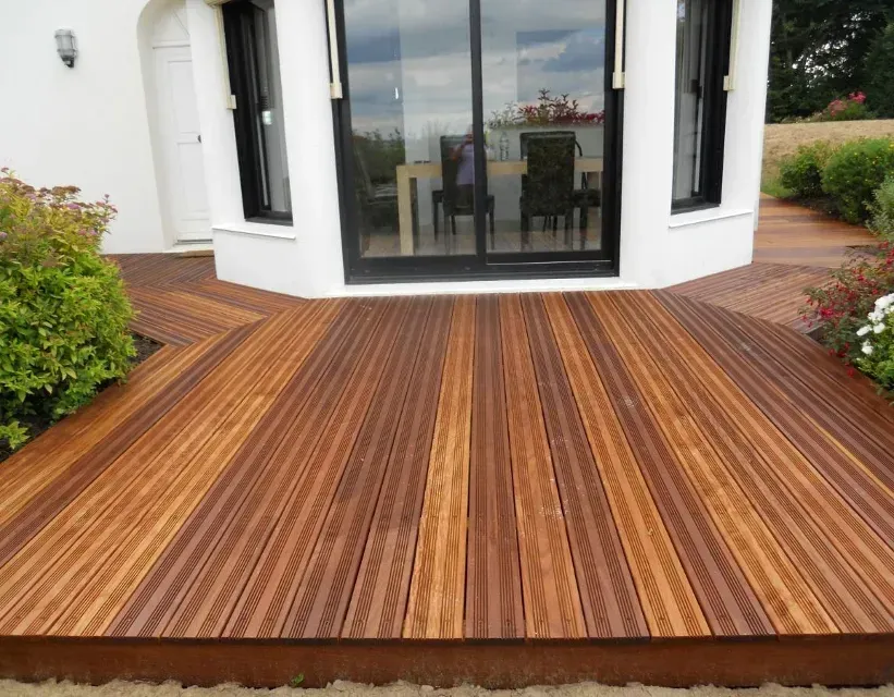 Terrasse en bois devant une maison blanche avec de grandes portes vitrées et entourée de verdure.
