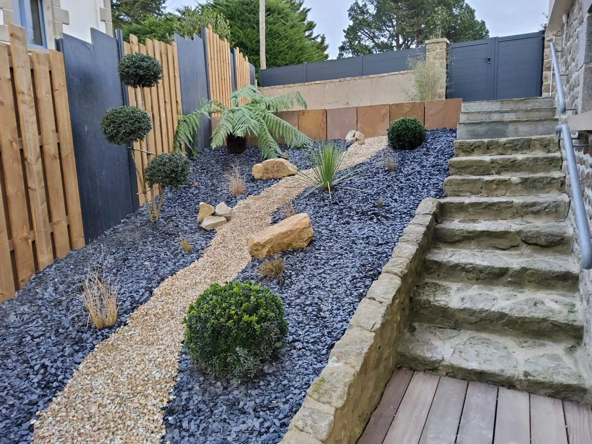 Cour arrière aménagée avec allée de gravier, plantes succulentes, clôture en bois et marches en béton.