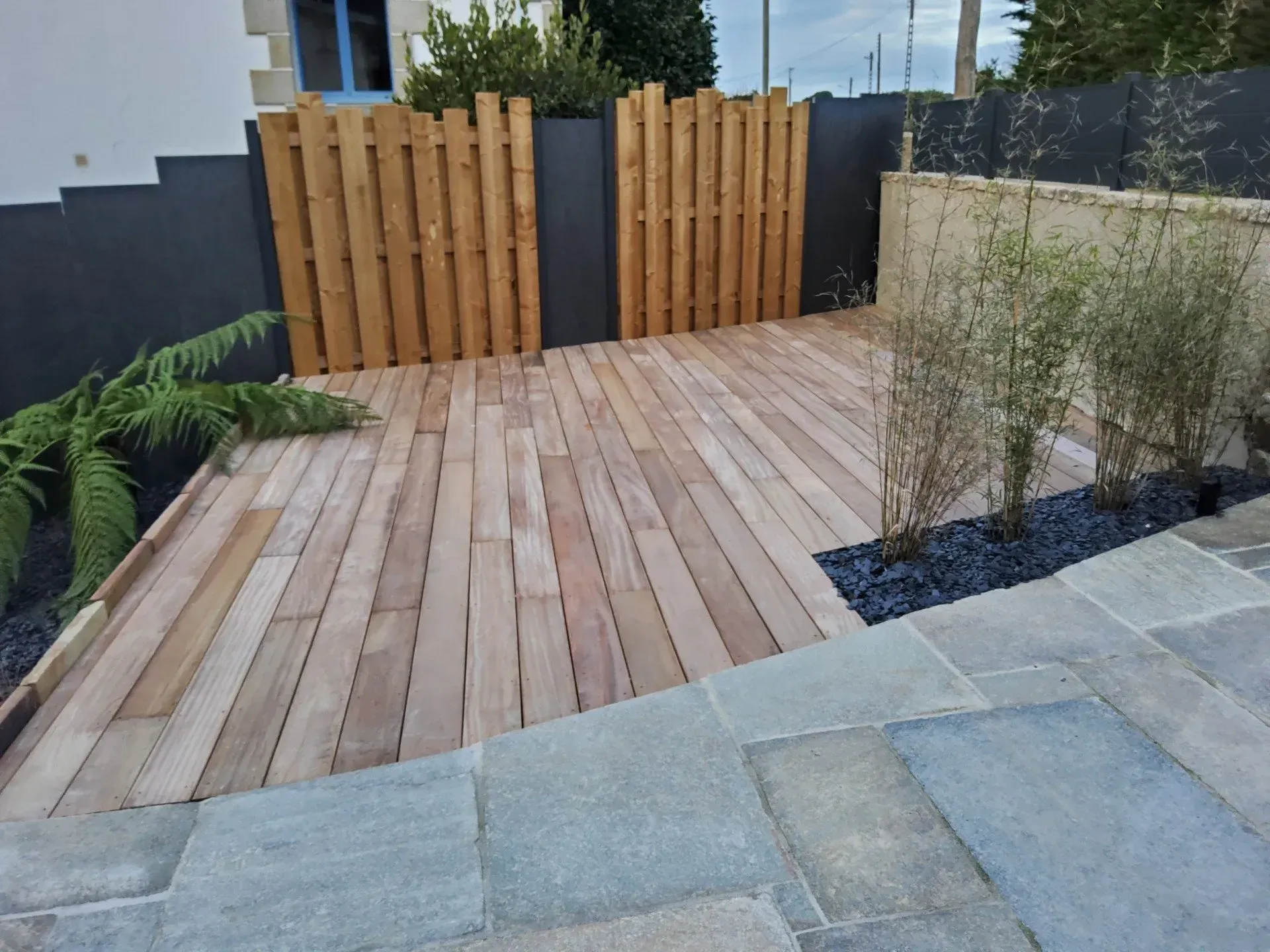 Terrasse moderne dans le jardin avec terrasse en bois, dalles de béton et jardinières en bambou menant à une clôture en bois.