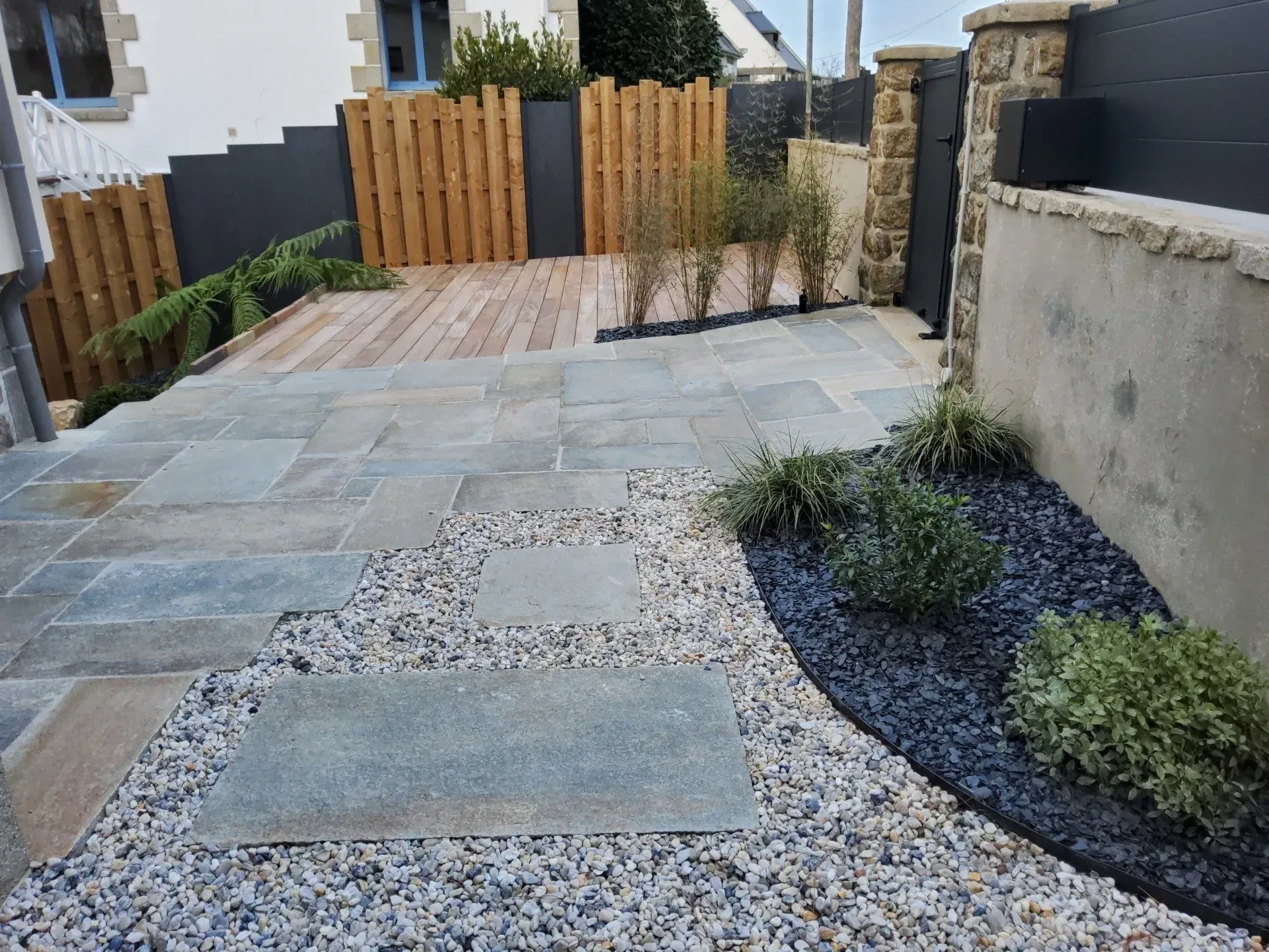Terrasse moderne dans le jardin avec pavage en pierre, bordure de gravier, clôture en bois et parterre de fleurs.