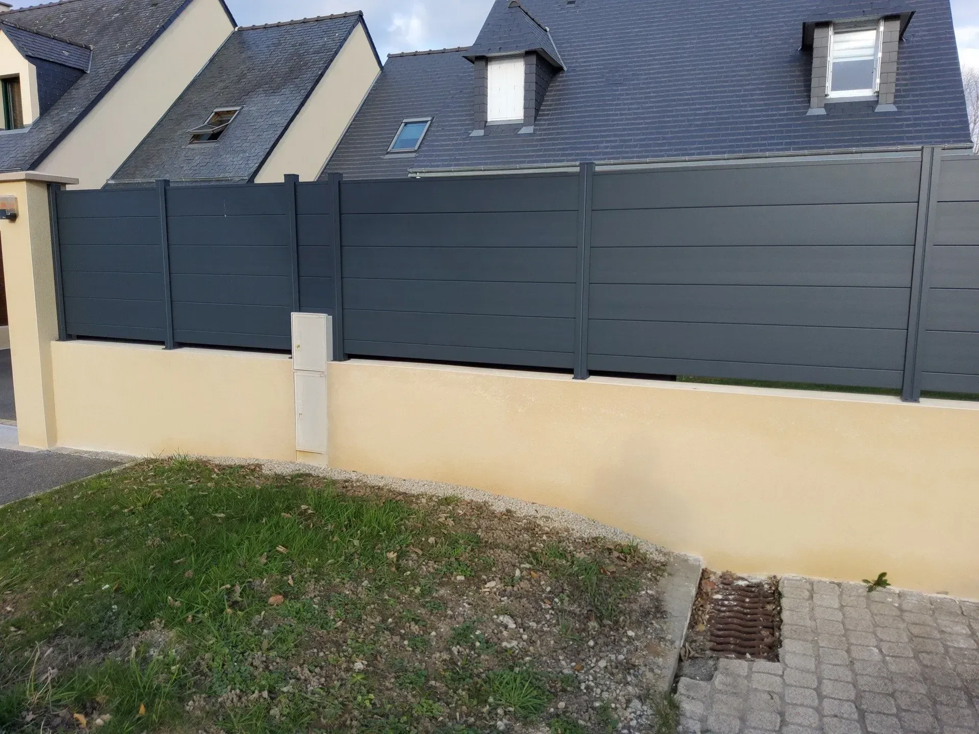 Une clôture gris foncé surmonte un mur jaune, bordant une pente herbeuse et une allée pavée devant les maisons.
