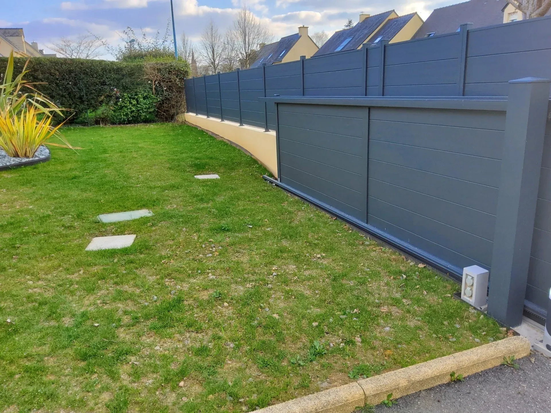 Pelouse arrière bordée d'une clôture et d'un portail bleu-gris sous un ciel partiellement nuageux.