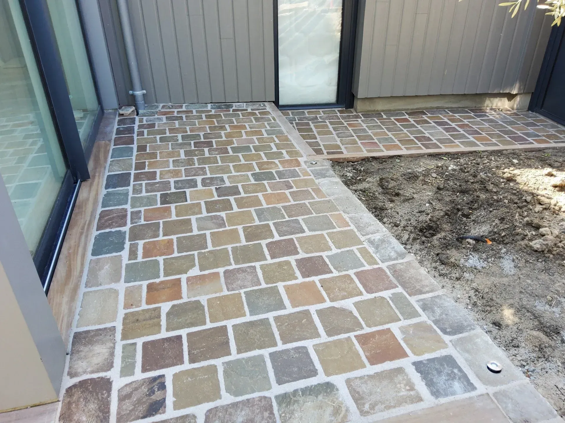 Terrasse pavée de briques à côté d'une porte vitrée, avec une zone de terre à droite et un bardage gris à l'arrière.