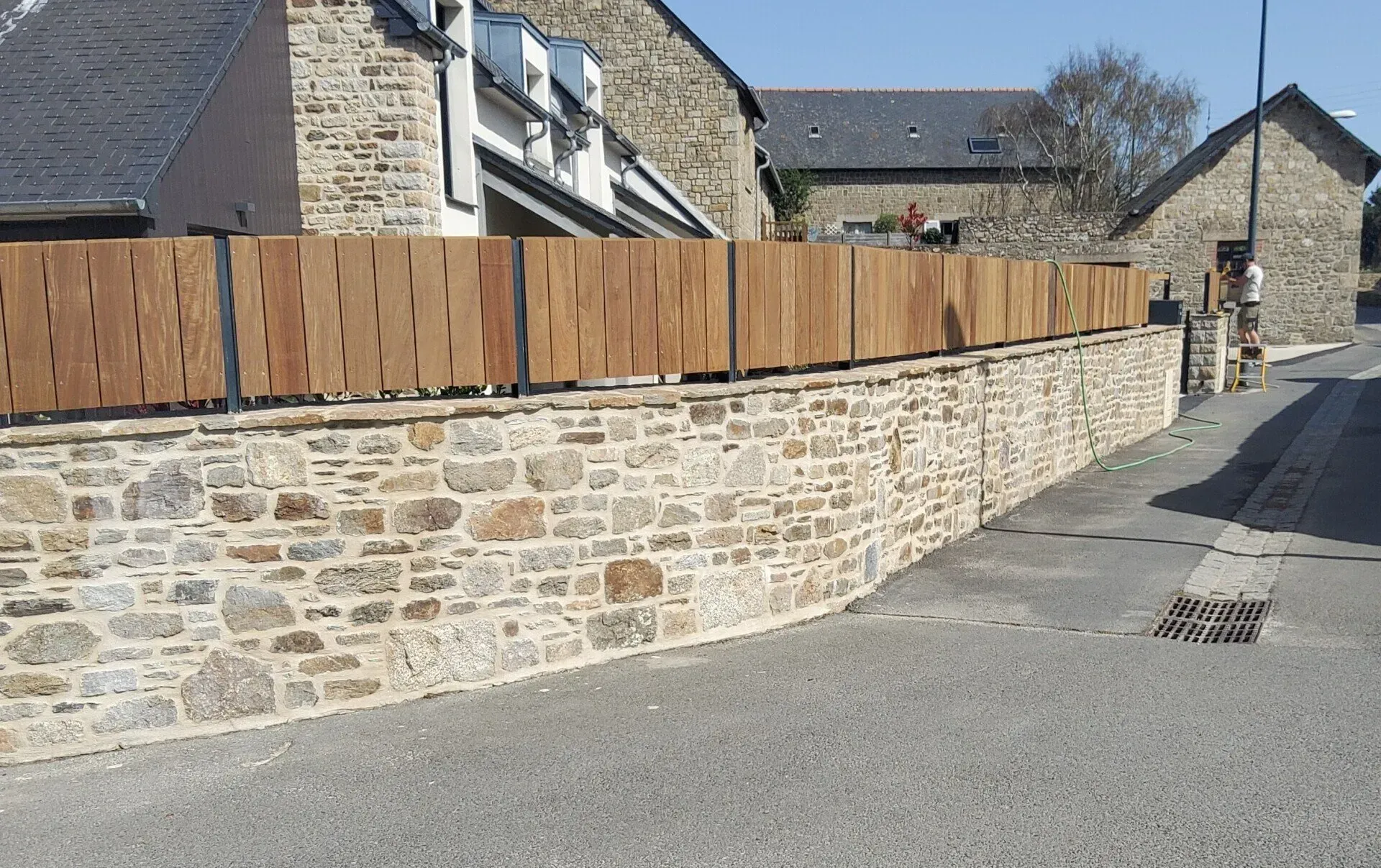 Mur en pierre incurvé avec une clôture en bois longeant une rue pavée étroite, bordée de maisons en pierre.