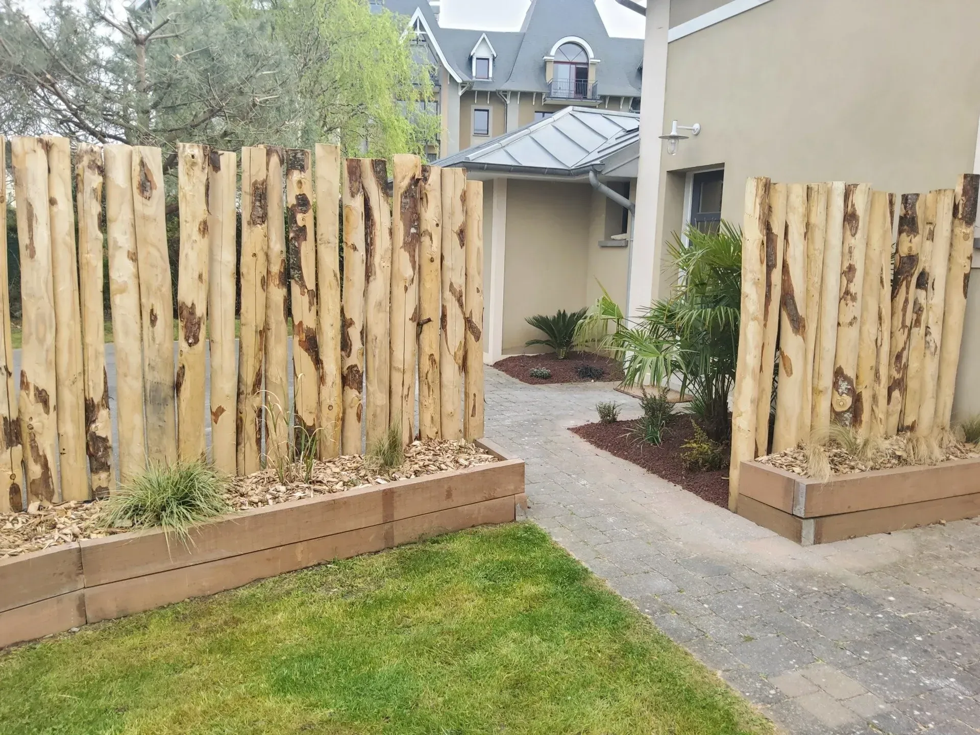 Des panneaux de clôture en bois bordent un chemin de jardin menant à une maison au toit gris et à la pelouse verte.