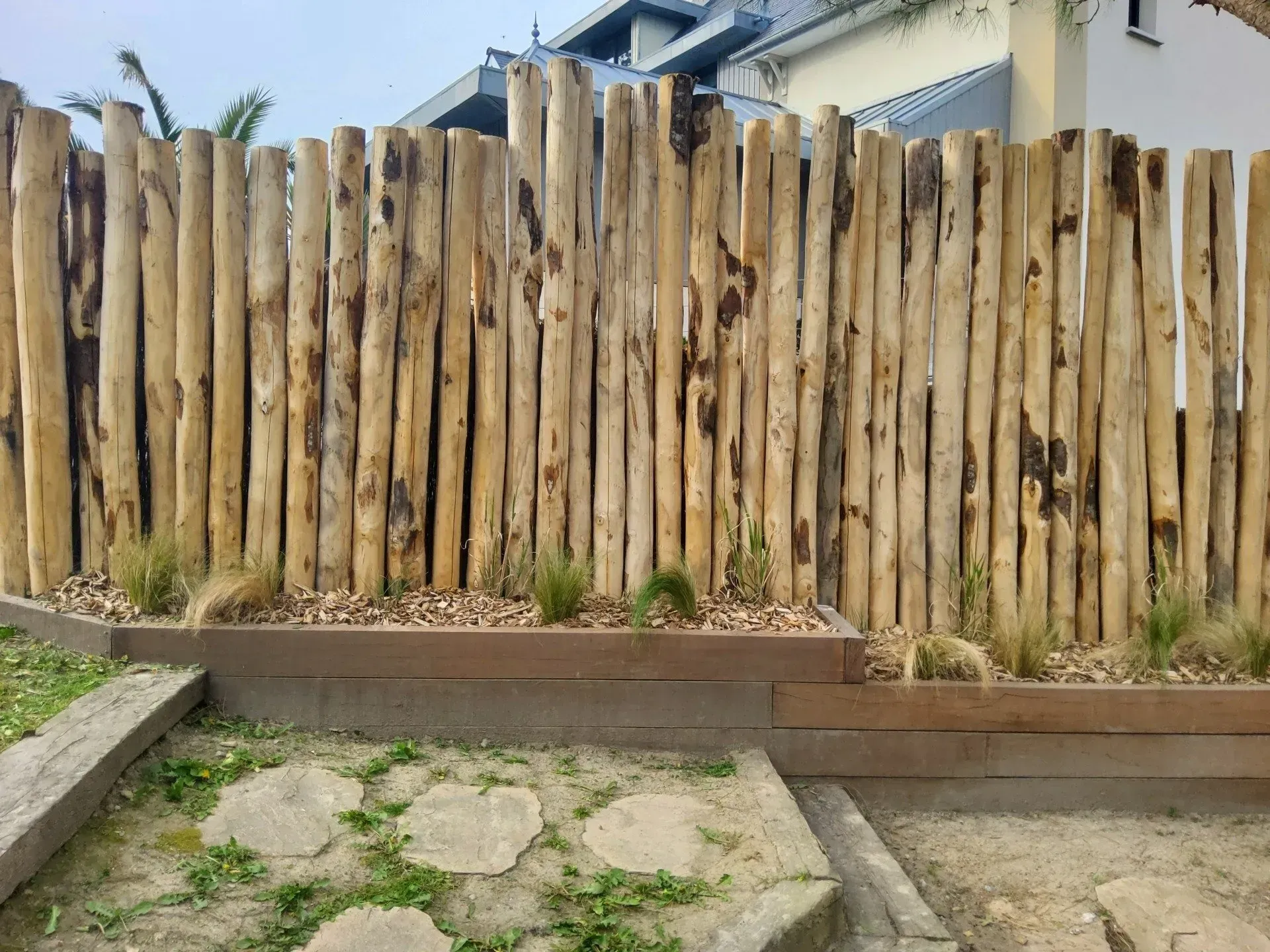 Clôture en bois patinée derrière un parterre surélevé et une terrasse en pierre avec quelques mauvaises herbes éparses.