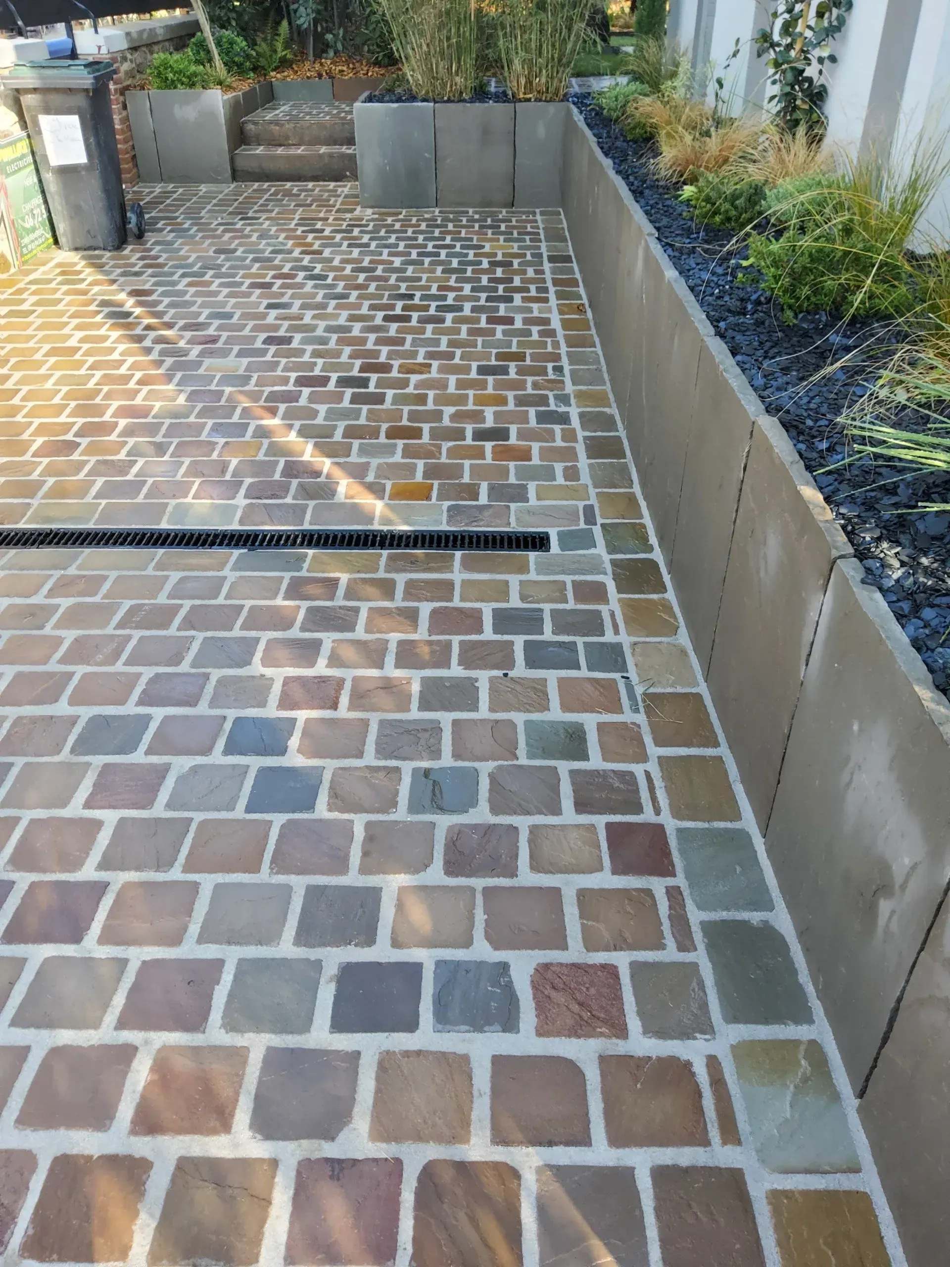 Allée pavée de briques avec une longue grille d'évacuation des eaux, bordée de jardinières surélevées en béton et de verdure.