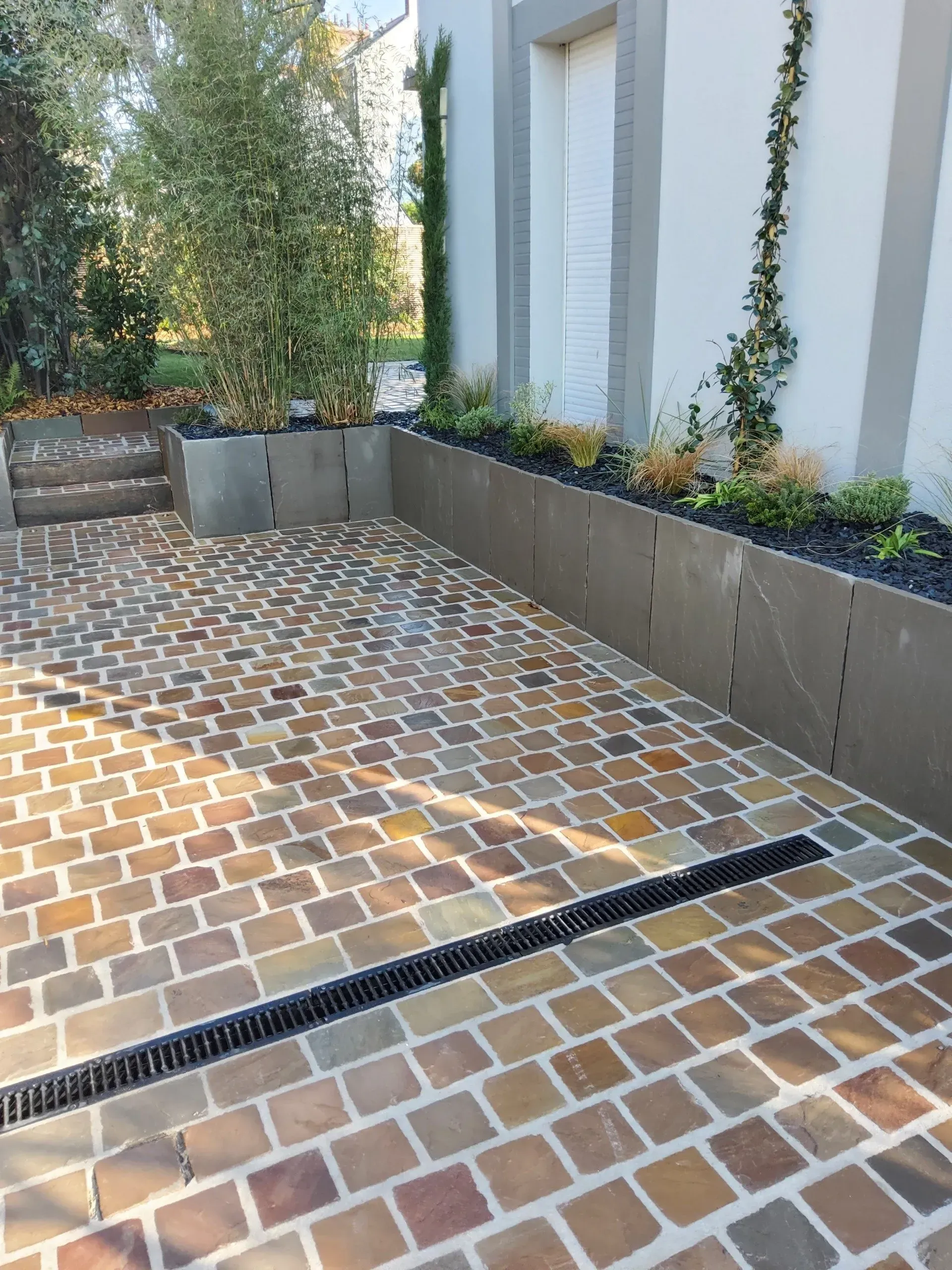 Terrasse extérieure avec pavés de briques beiges, jardinières surélevées et grille d'évacuation le long d'un mur blanc.