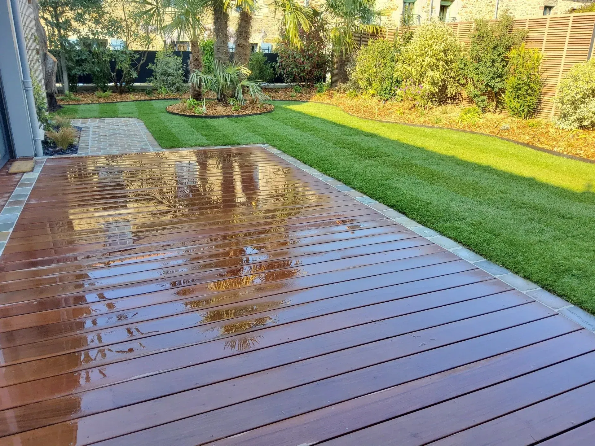 Terrasse en bois mouillée jouxtant une pelouse verte et un jardin paysager à l'arrière.