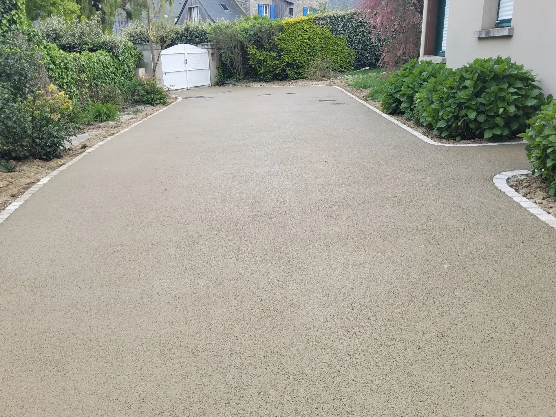 Allée large en gravier beige menant à un garage blanc, bordée d'arbustes verts et d'une maison sur la droite.