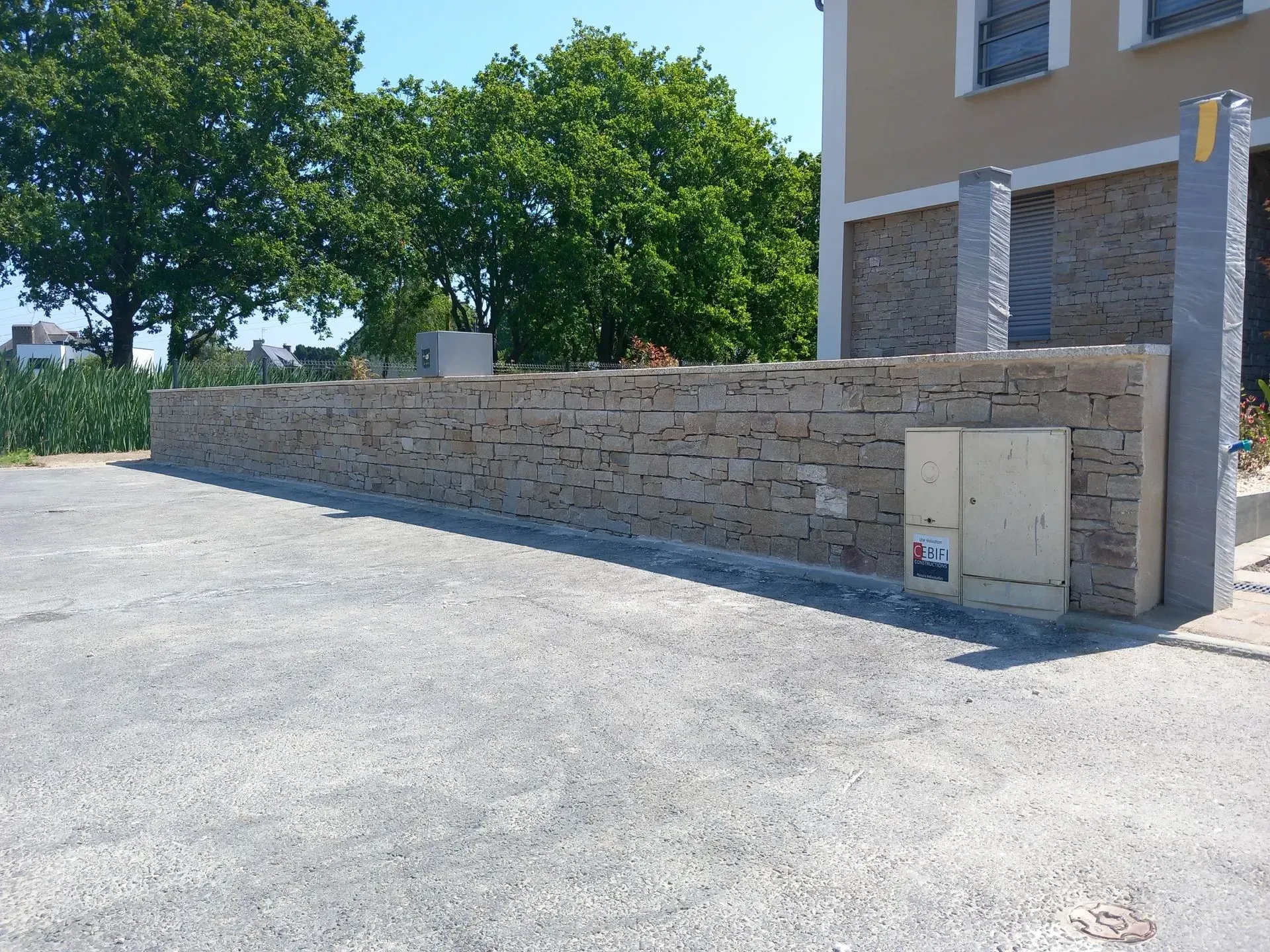 Mur de pierre le long d'une allée de gravier, à côté d'un bâtiment beige et d'arbres, en plein jour.