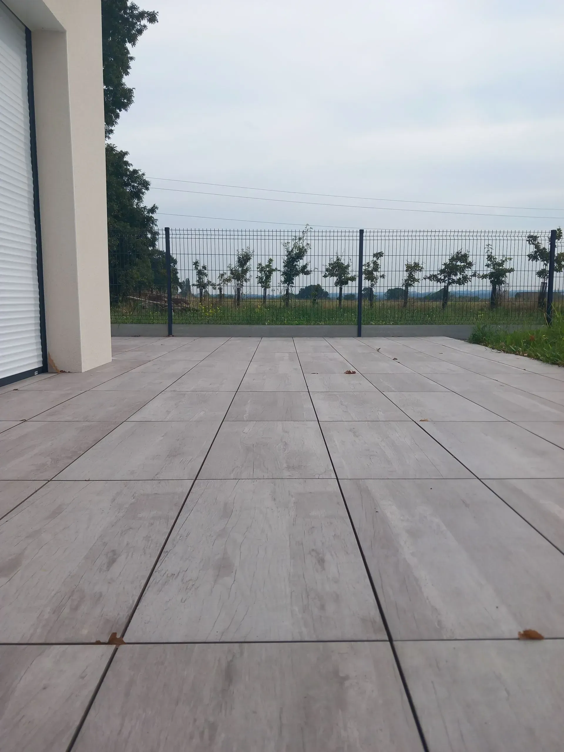 Patio carrelé vide à côté d'un bâtiment, avec une clôture et des arbres au loin sous un ciel nuageux.