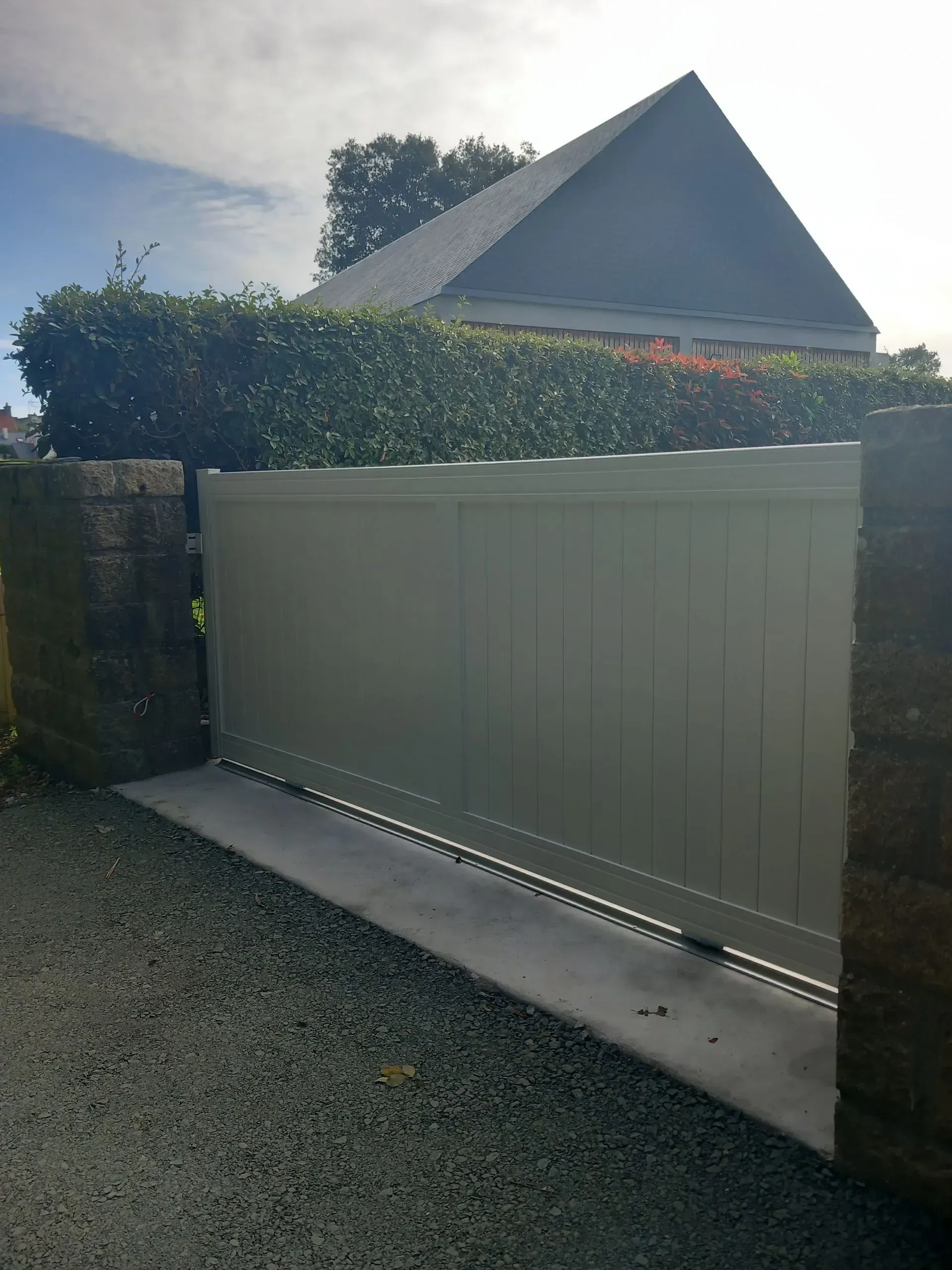 Portail d'allée gris clair fermé, entre des piliers en briques, avec une haie et le toit d'une maison derrière.