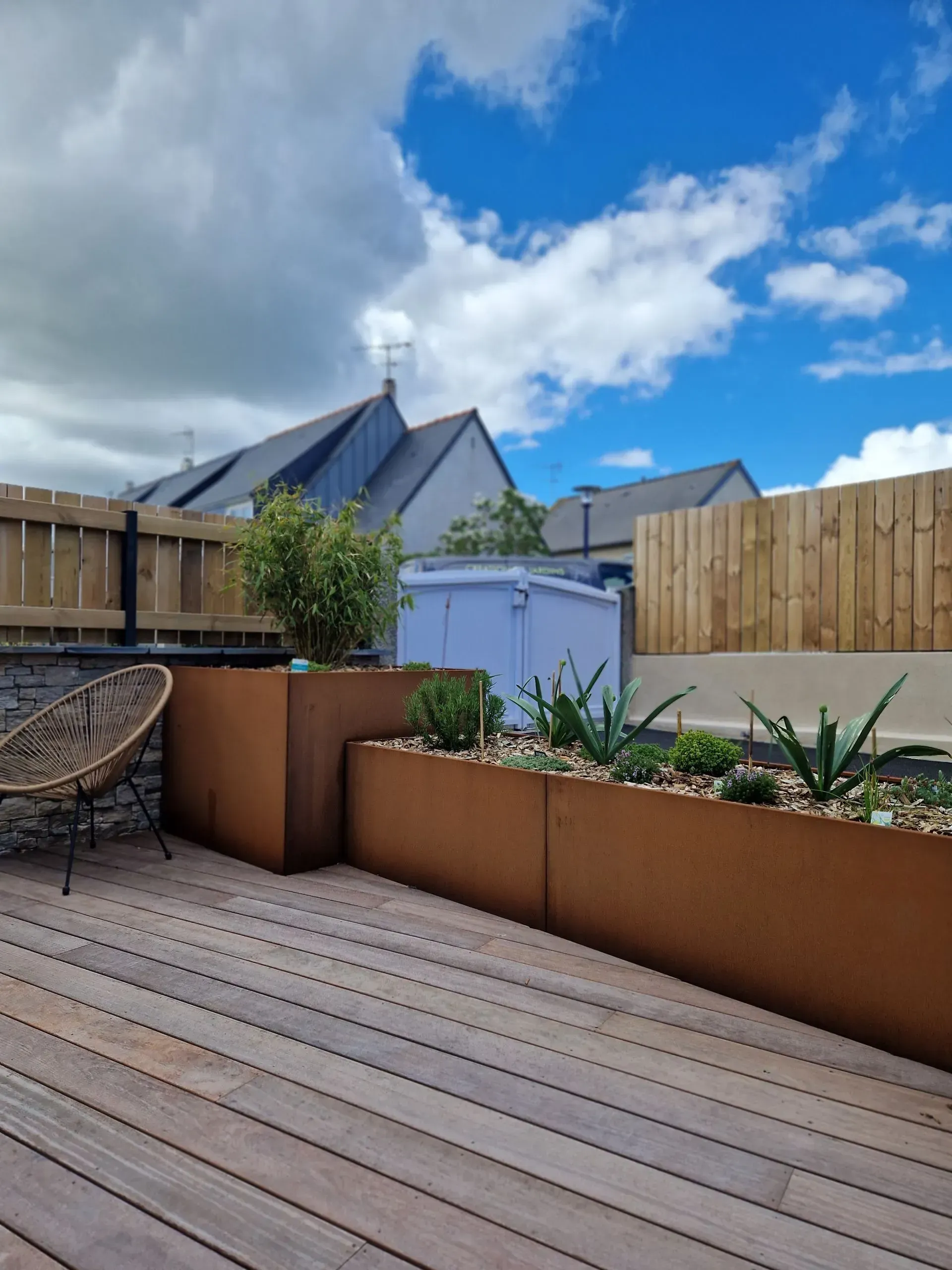 Terrasse en bois avec jardinières beiges, chaise en osier et ciel nuageux au-dessus des toits voisins.